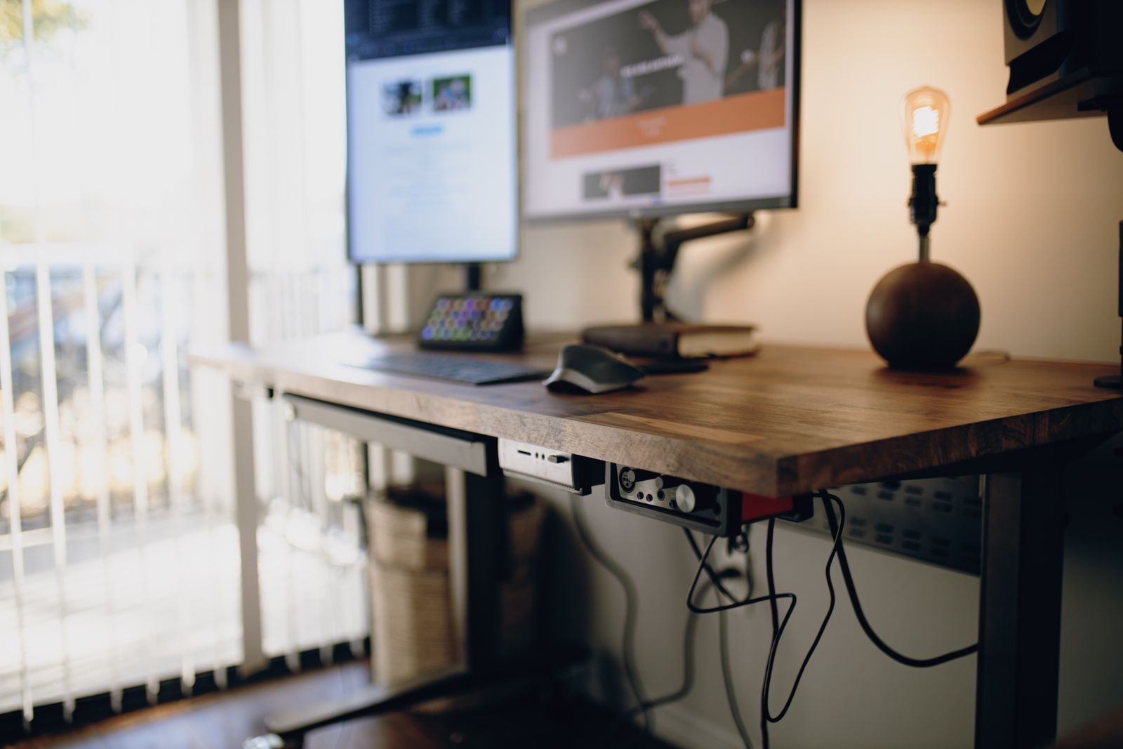 A height-adjustable standing desk with a wooden top and monitors, set up by a window in a bright room.
